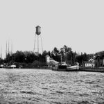 Seen in 1908, the Life-Saving station was situated below and to the west of the water tower which went up in 1906. At far right was Charlevoix’s second electric plant. Half of this building, the north part shown here, was removed to make way for the second fish hatchery in 1917. The photo was taken from the north pier. At far left by the trestle appears the 1890 storage building for the oil used in the lighthouse. Just to the right of the three-masted schooner’s stern appears a little curved white structure on the revetment in front of the station Life-Saving Station 1908