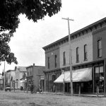 Bridge Street circa 1890, looking south from Park Avenue. The Bartholomew block, as this structure was known, held an auditorium spread across the top floor that seated six hundred. Two men stand in the doorway of the Frank Wood Barber Shop that provided bathing facilities upstairs. Men could have “Whisker Dyeing Done in Artistic Style” here. The Charlevoix Cigar Company shared the upstairs premises. Its horizontal sign caps the barber pole. The entire building block was destroyed by fire in the winter of 1923. The auditorium was not rebuilt. Bridge Street 1890