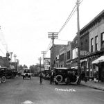 Bridge Street, 1922, looking south at the height of the summer season, also from Park Avenue. The hardware store has become Fessenden’s Drugstore. The Drugs/Books sign in the middle is on the tall wooden Clinton Street wall of the Central Drug Store. Parking then, as now, was a headache as the latest models swarmed into town, many of them brought aboard passenger liners rather than risk damage on terrible roads. Another traffic directional “button” appears in the pavement at bottom. Bridge Street 1922